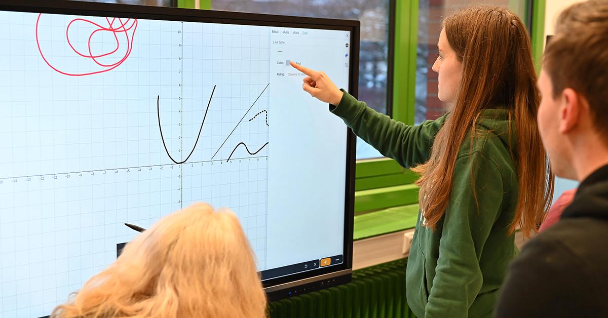 Eine Frau zeigt auf einen Bildschirm mit mathematischen Graphen. | A woman points at a screen showing mathematical graphs.